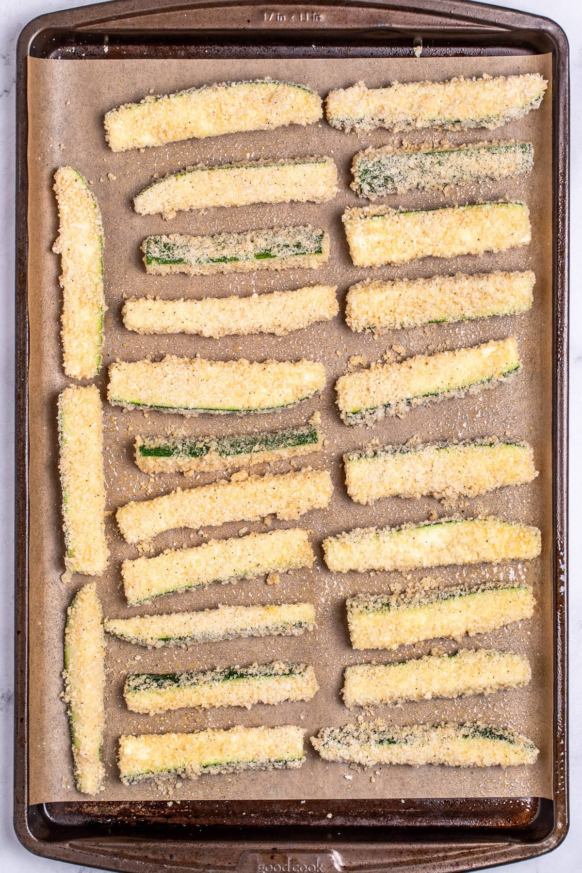 A sheet pan lined with parchment paper, with zucchini strips breaded and ready to be baked.