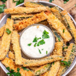A bowl of baked zucchini fries with a smaller bowl of dipping sauce in the middle topped with fresh chopped parsley.