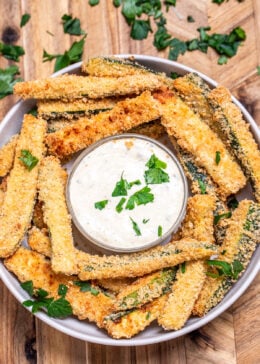 A bowl of baked zucchini fries with a smaller bowl of dipping sauce in the middle topped with fresh chopped parsley.