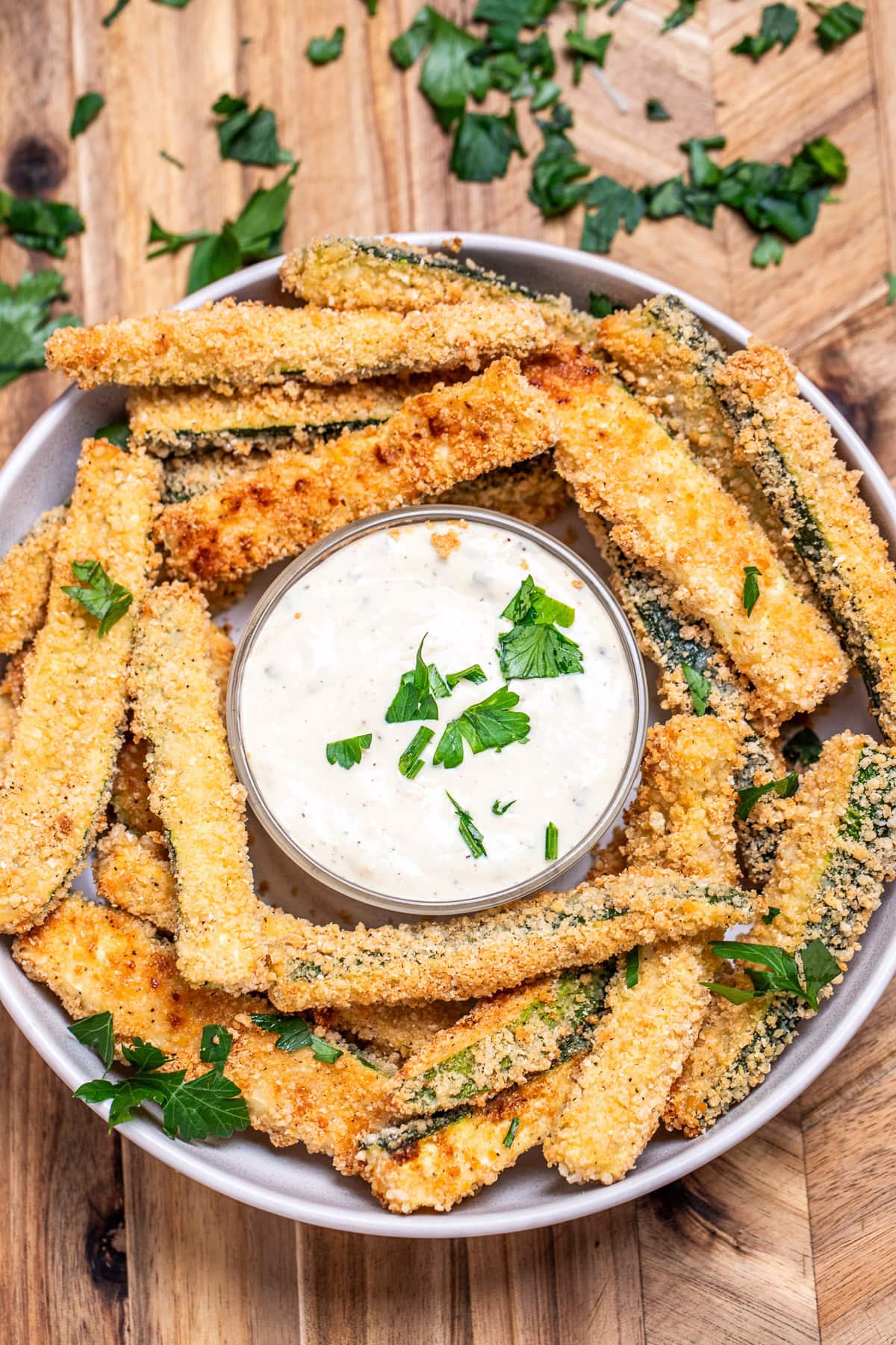 A bowl of baked zucchini fries with a smaller bowl of dipping sauce in the middle topped with fresh chopped parsley.