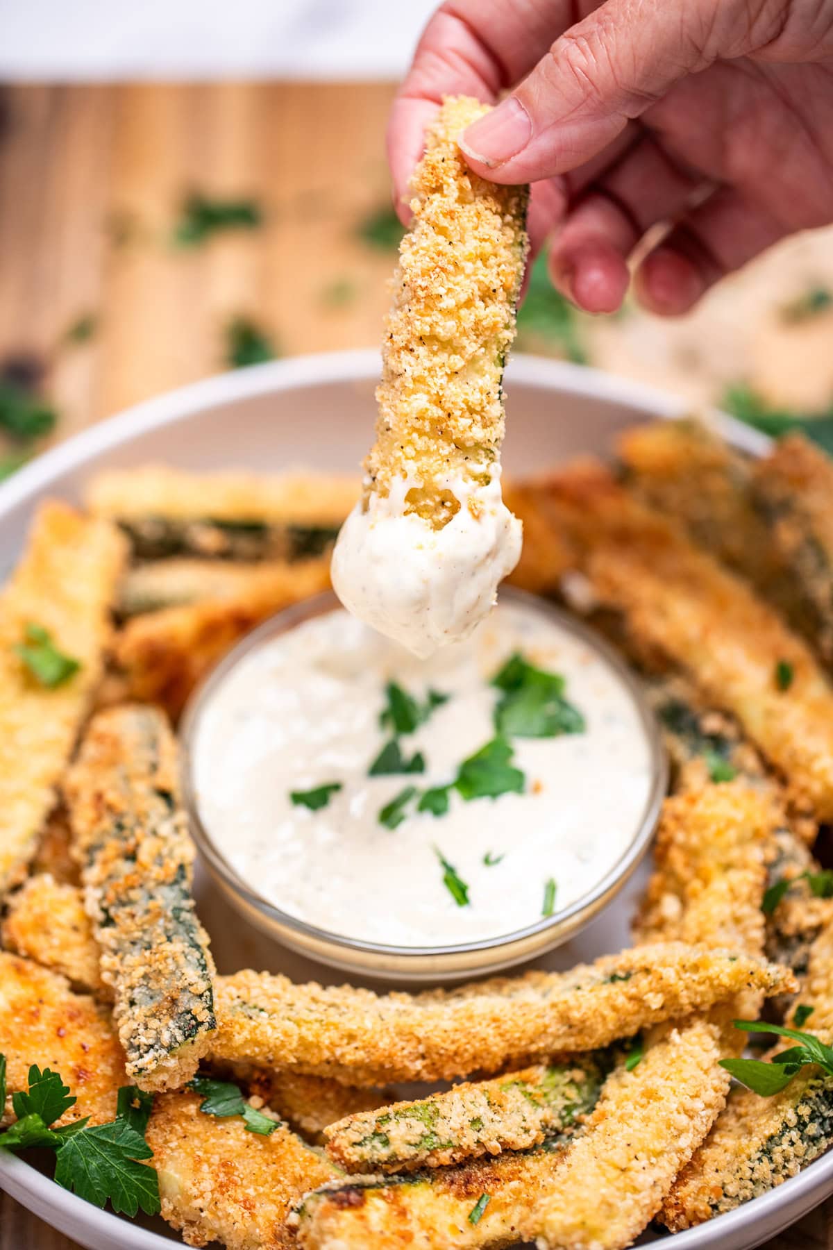 A bowl of baked zucchini fries with a smaller bowl of dipping sauce in the middle topped with fresh chopped parsley, with a hand dipping a fry into the sauce.