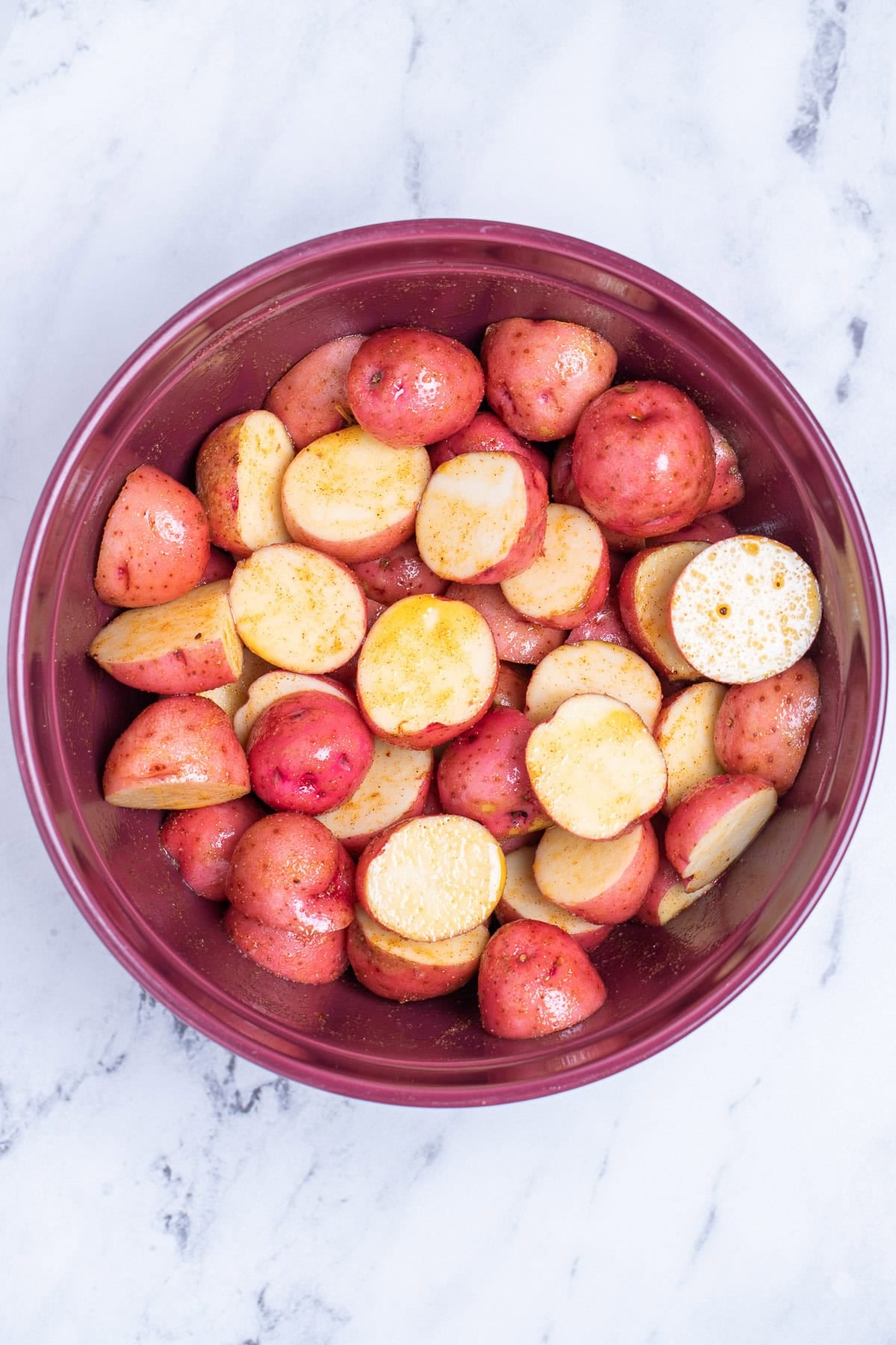 A red bowl full of halved red potatoes tossed with olive oil and seasoning before being cooked.