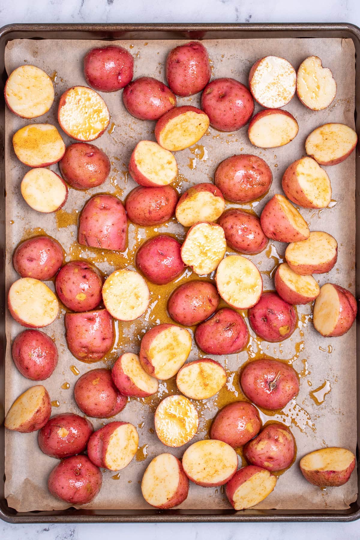 A sheet pan lined with parchment paper with halved baby red potatoes coated in olive oil and seasoning, before being roasted.