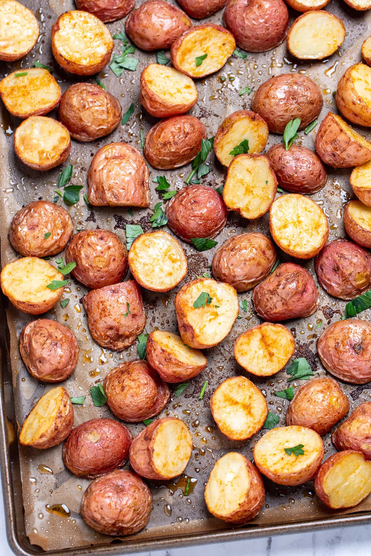 A sheet pan lined with parchment paper, with seasoned halved red potatoes after being roasted, topped with chopped fresh parsley.
