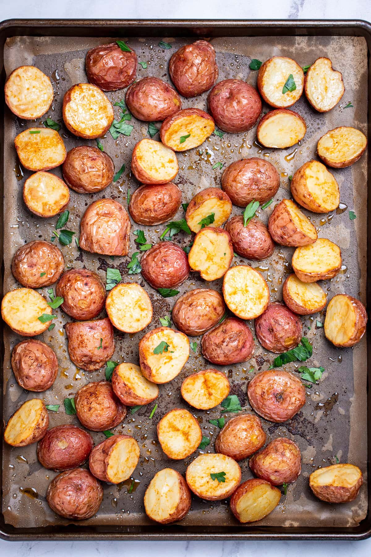 A sheet pan lined with parchment paper, with seasoned halved red potatoes after being roasted, topped with chopped fresh parsley.