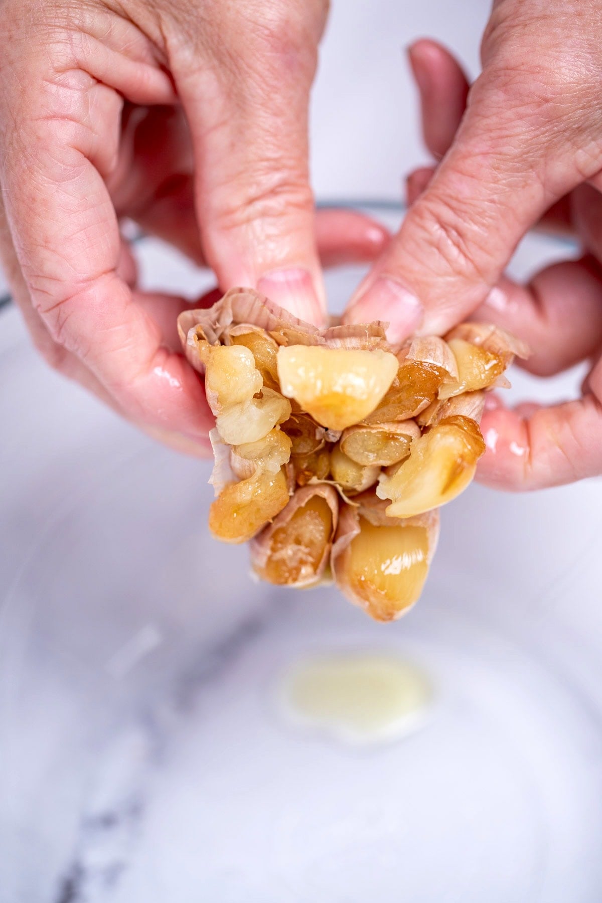 Two hands squeezing the soft garlic bulbs out of a roasted head of garlic above a bowl.