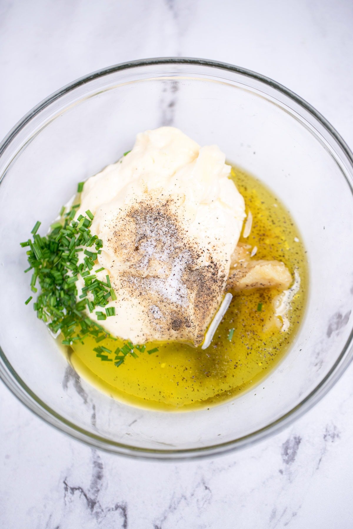 A glass bowl on a table, full of individual ingredients for roasted garlic aioli before being mixed together.