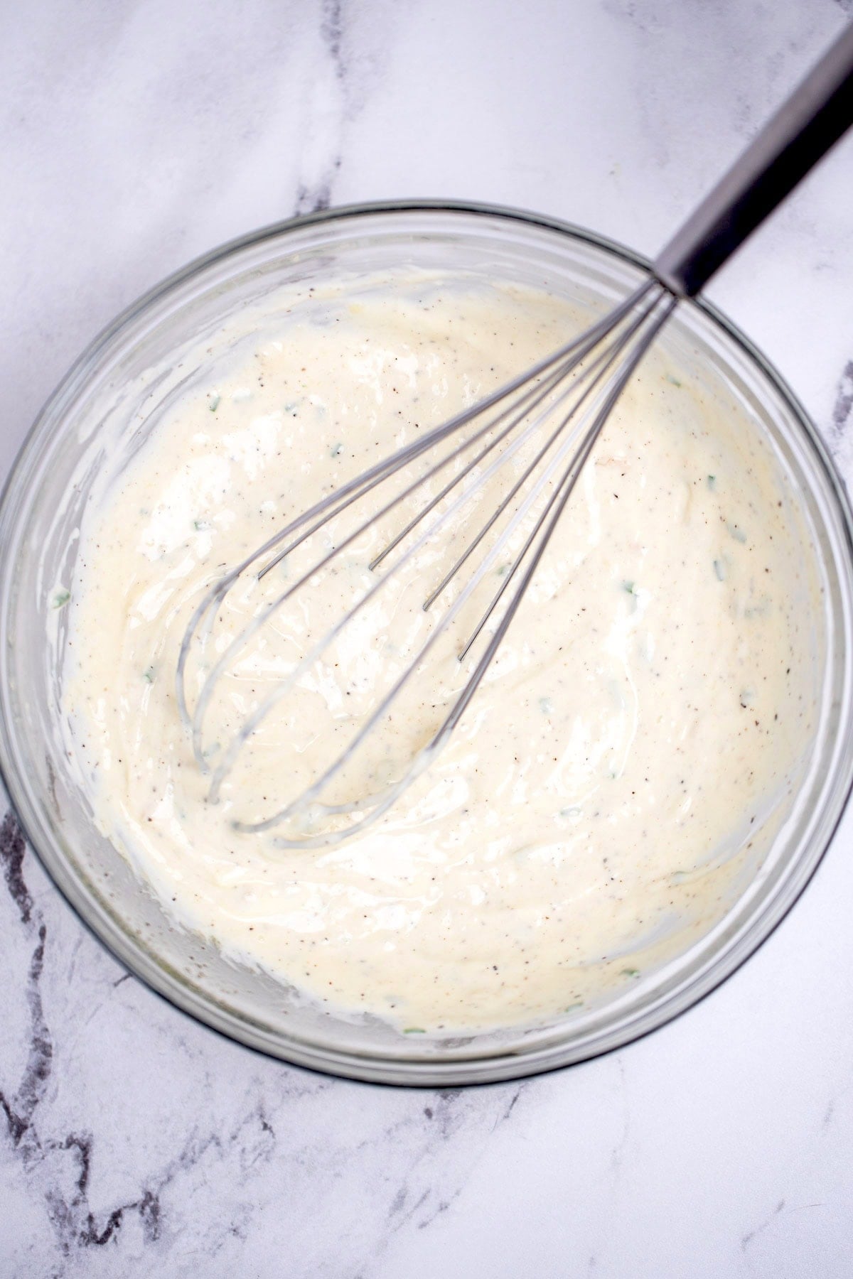 A glass bowl on a table with finished roasted garlic aioli, and a whisk resting in the sauce.