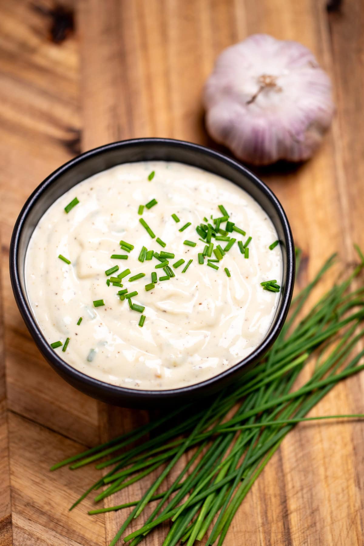 A small bowl of roasted garlic aioli topped with fresh chives, on a cutting board next to more chives and a bulb of garlic.