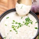 A small bowl of roasted garlic aioli topped with fresh chives, on a cutting board next to more chives and a bulb of garlic. A spoon is dipping into the sauce and lifting some up.