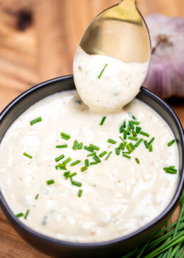 A small bowl of roasted garlic aioli topped with fresh chives, on a cutting board next to more chives and a bulb of garlic. A spoon is dipping into the sauce and lifting some up.