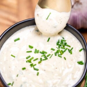A small bowl of roasted garlic aioli topped with fresh chives, on a cutting board next to more chives and a bulb of garlic. A spoon is dipping into the sauce and lifting some up.