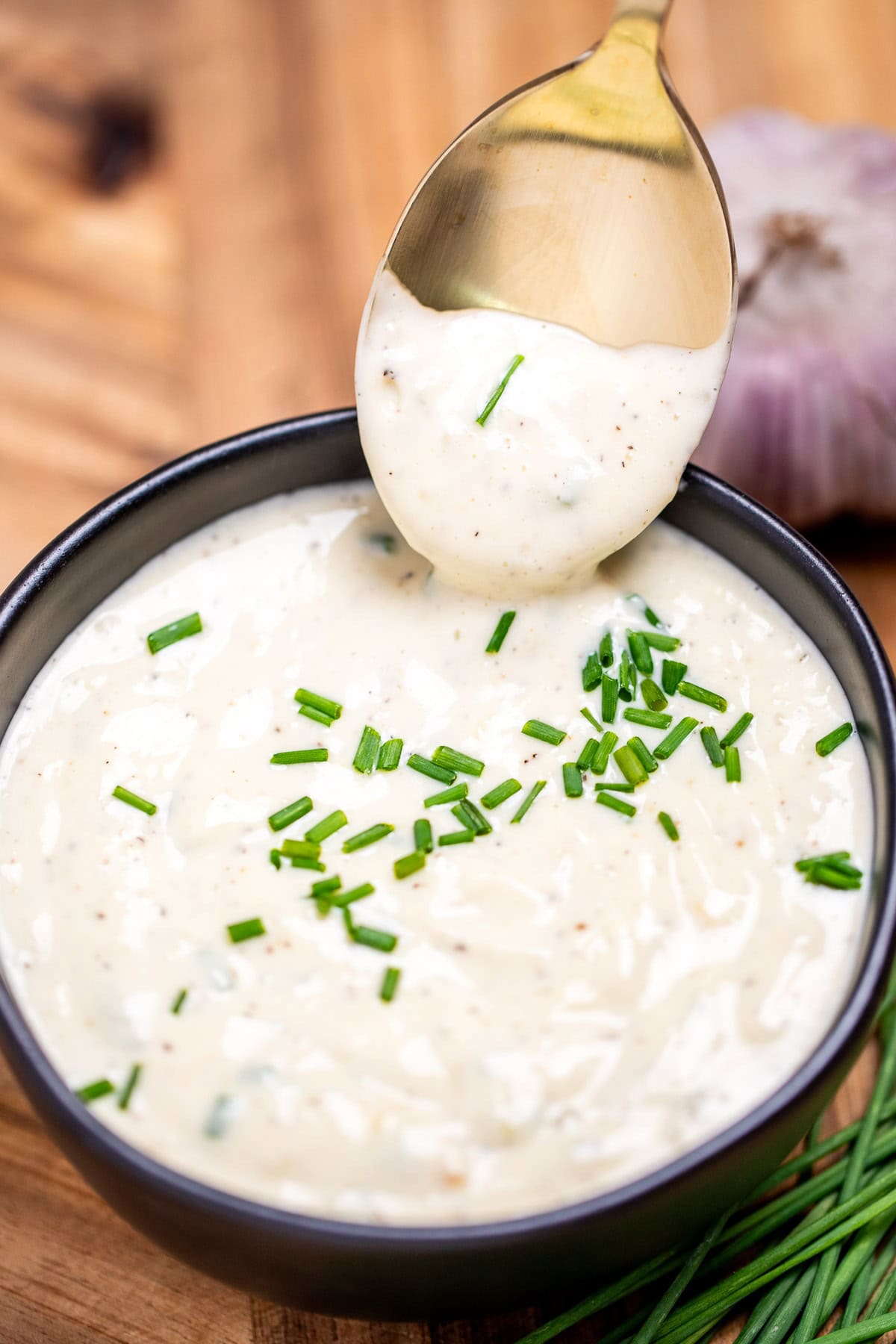 A small bowl of roasted garlic aioli topped with fresh chives, on a cutting board next to more chives and a bulb of garlic. A spoon is dipping into the sauce and lifting some up.