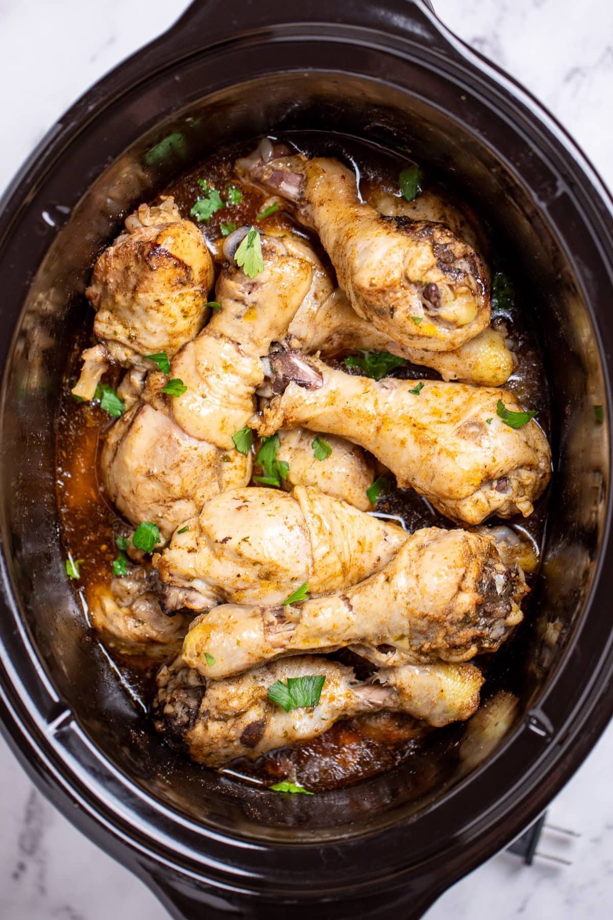A slow cooker full of cooked chicken drumsticks in broth, topped with fresh parsley.