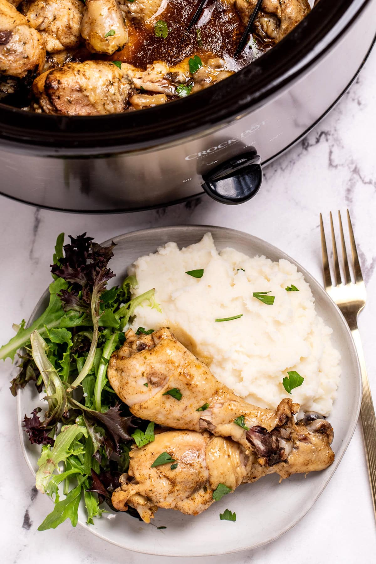 A plate with two chicken legs next to salad and mashed potatoes, in front of a slow cooker.