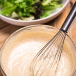A bowl of lemon tahini dressing with a whisk resting in it, in front of a bowl of salad with lettuce, cherry tomatoes, and chickpeas.