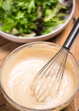 A bowl of lemon tahini dressing with a whisk resting in it, in front of a bowl of salad with lettuce, cherry tomatoes, and chickpeas.