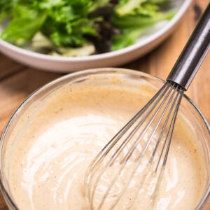 A bowl of lemon tahini dressing with a whisk resting in it, in front of a bowl of salad with lettuce, cherry tomatoes, and chickpeas.