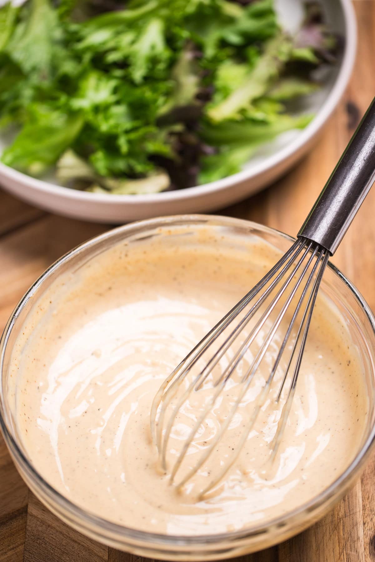 A bowl of lemon tahini dressing with a whisk resting in it, in front of a bowl of salad with lettuce, cherry tomatoes, and chickpeas.