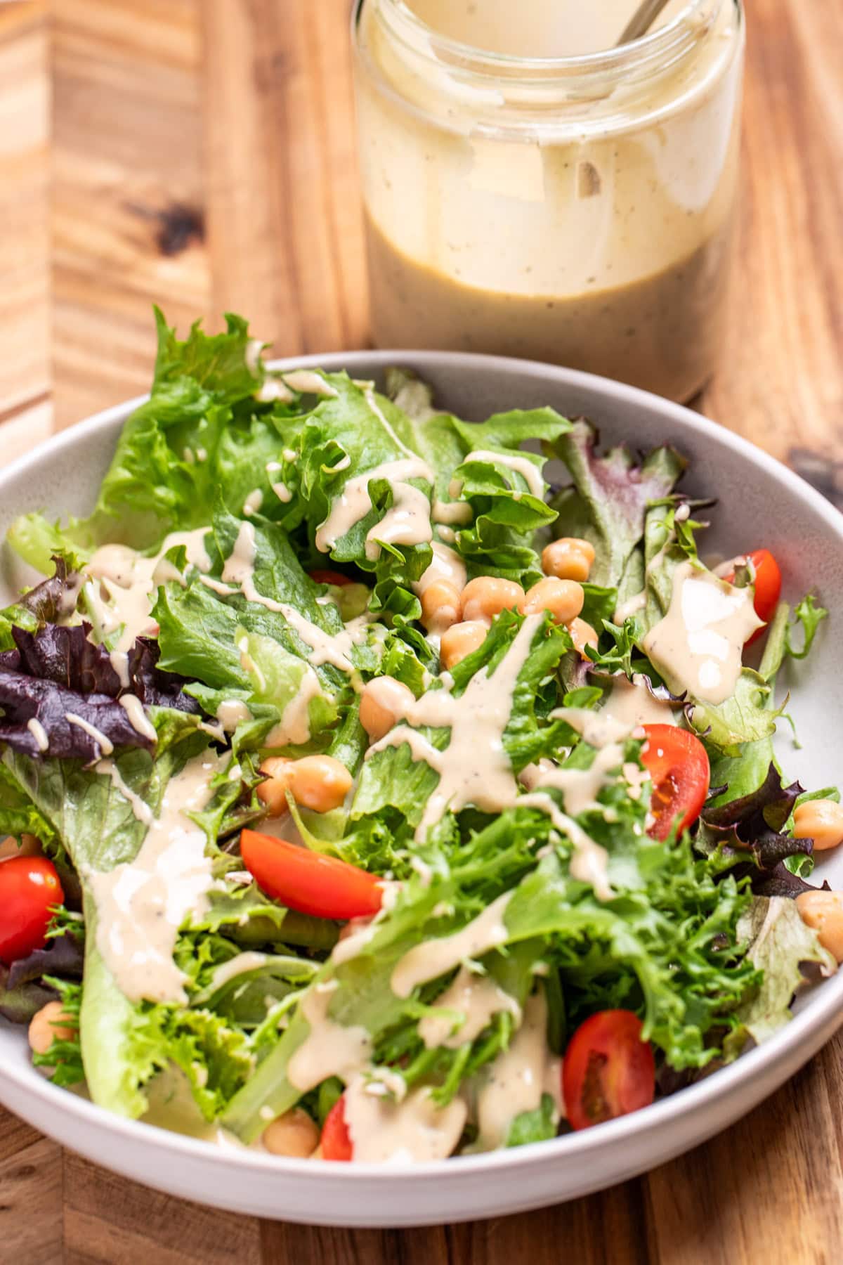 A salad with lettuce, cherry tomatoes, and chickpeas topped with lemon tahini dressing in a bowl on a cutting board, in front of a jar of dressing.