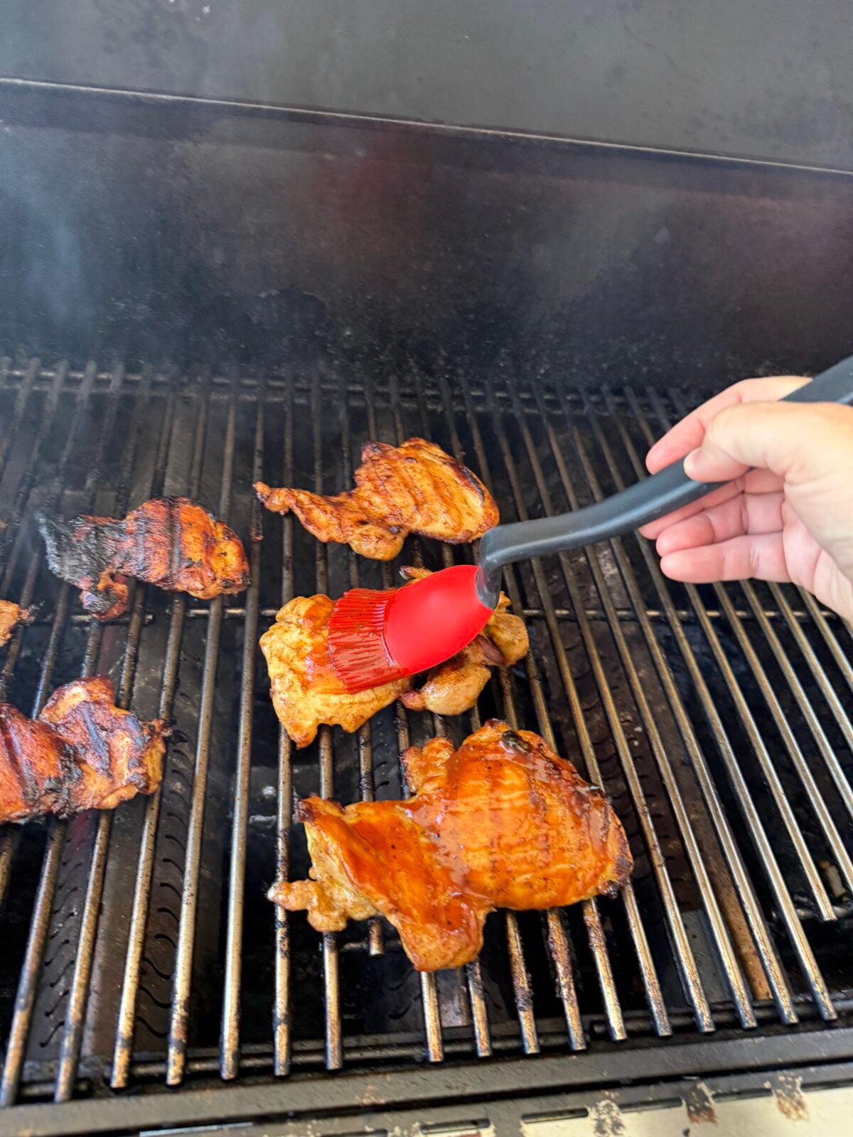 A hand using a sauce brush to brush BBQ sauce on top of chicken thighs while cooking on the grill.