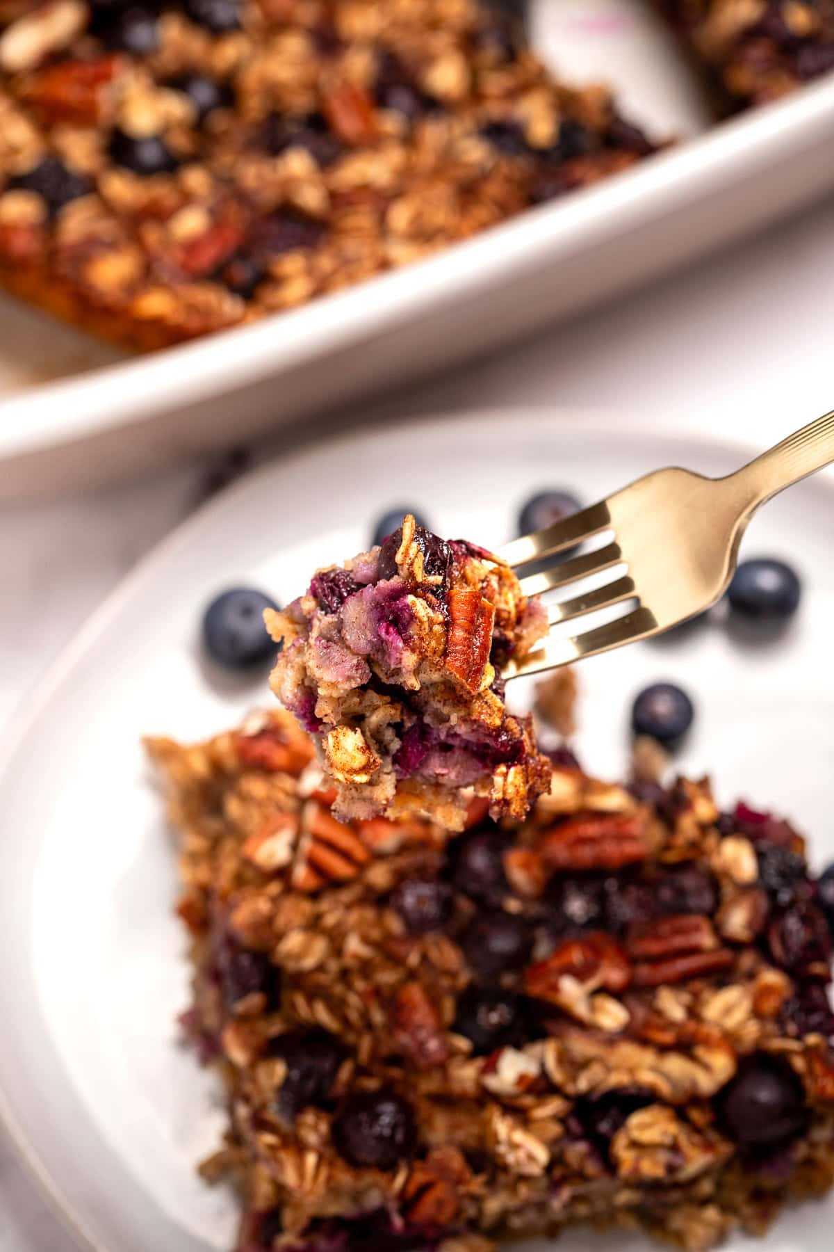 A plate with a square of blueberry baked oatmeal next to some fresh blueberries, in front of a baking dish of oatmeal, and a fork lifting some up.
