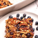 A plate with a square of blueberry baked oatmeal next to some fresh blueberries, in front of a baking dish of oatmeal.