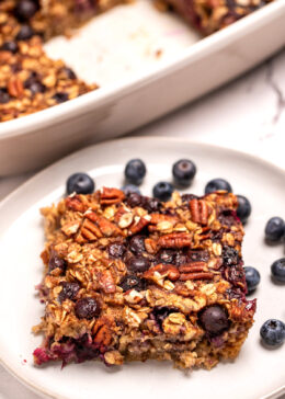 A plate with a square of blueberry baked oatmeal next to some fresh blueberries, in front of a baking dish of oatmeal.
