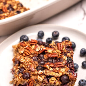 A plate with a square of blueberry baked oatmeal next to some fresh blueberries, in front of a baking dish of oatmeal.