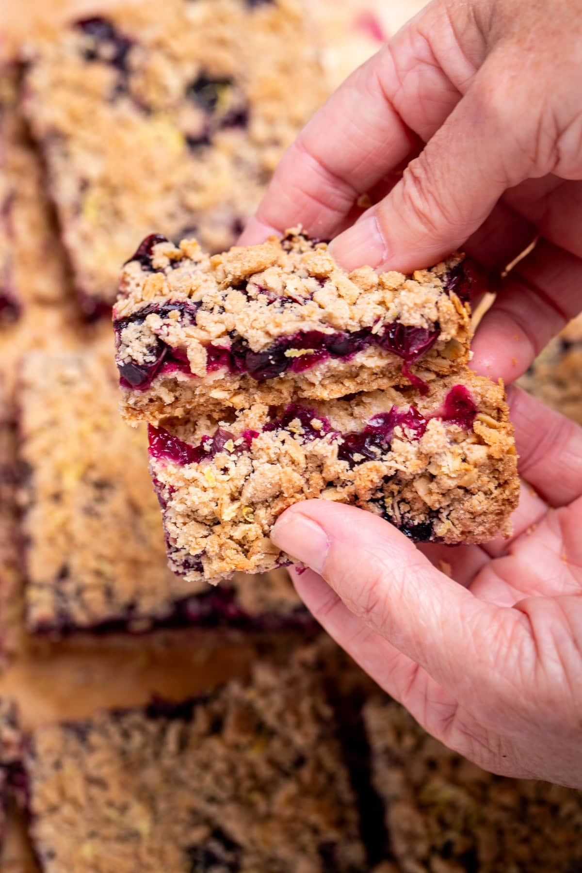 Two hands breaking a blueberry oatmeal bar in half to show the middle blueberry layer, above more bars.