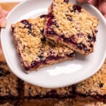 Two hands holding a plate with two blueberry oatmeal bars on it, above a pan of more bars.