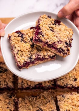 Two hands holding a plate with two blueberry oatmeal bars on it, above a pan of more bars.