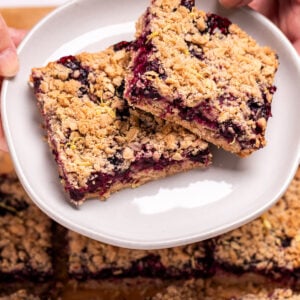 Two hands holding a plate with two blueberry oatmeal bars on it, above a pan of more bars.