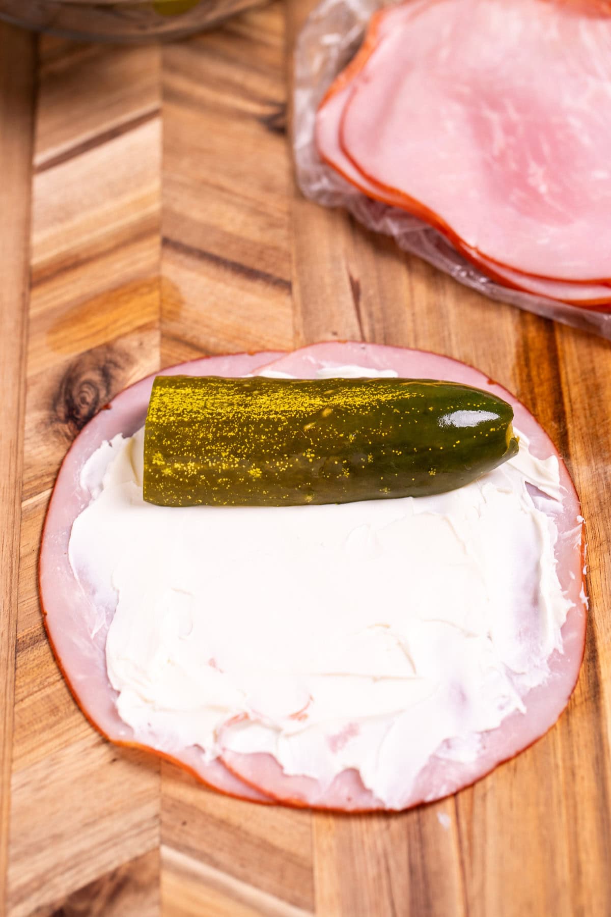 A piece of deli ham with cream cheese and a pickle, with more ham in the background.