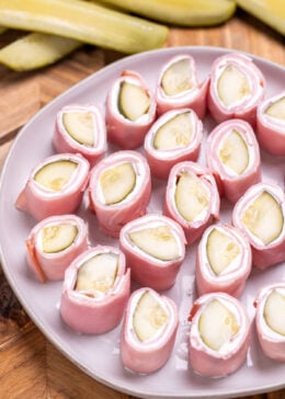 A plate resting on a cutting board with pieces of dill pickle roll ups ready to be served, with pickle spears next to it.