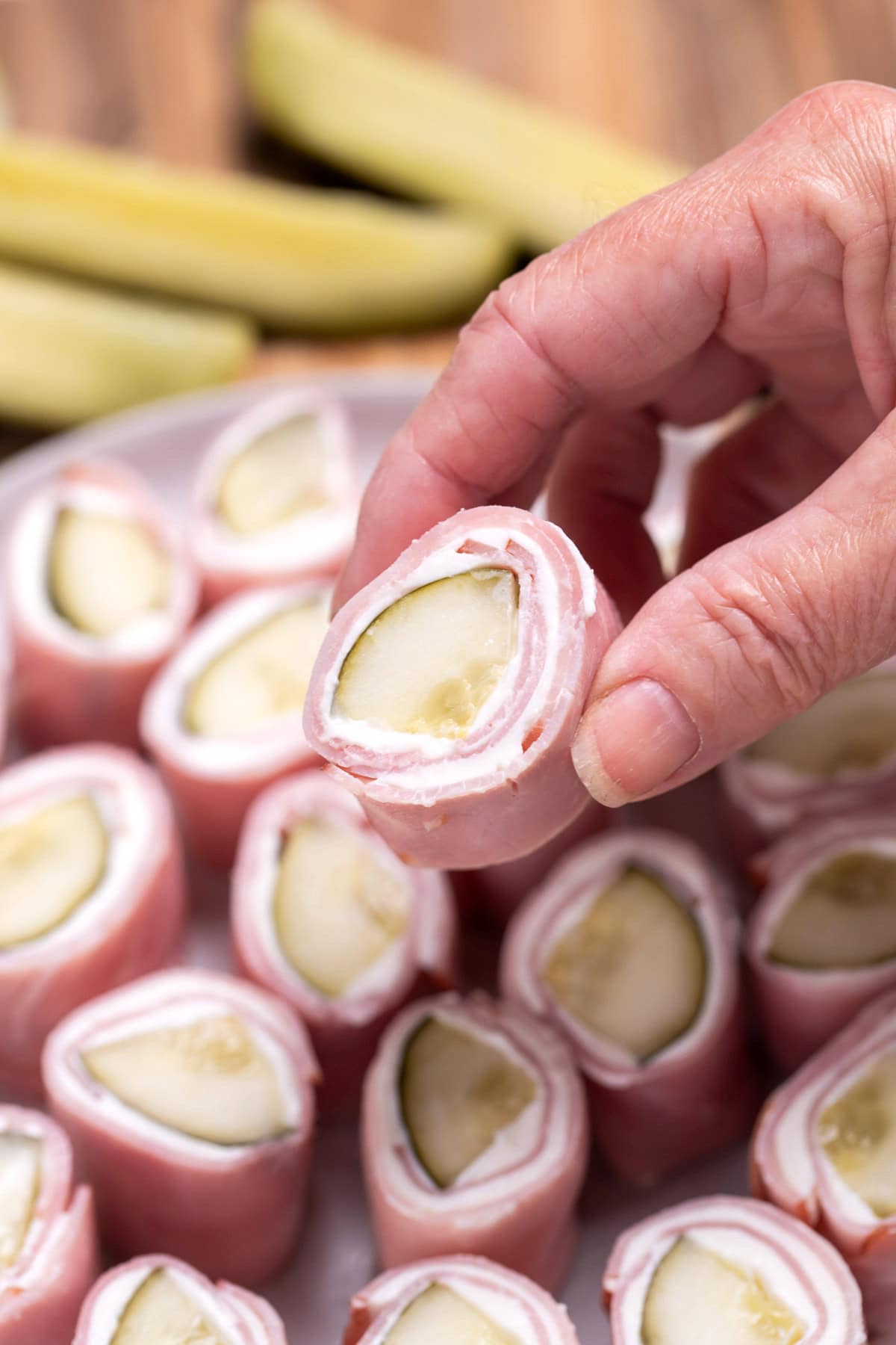 A plate resting on a cutting board with pieces of dill pickle roll ups ready to be served, with pickle spears next to it. A hand is lifting a piece up.