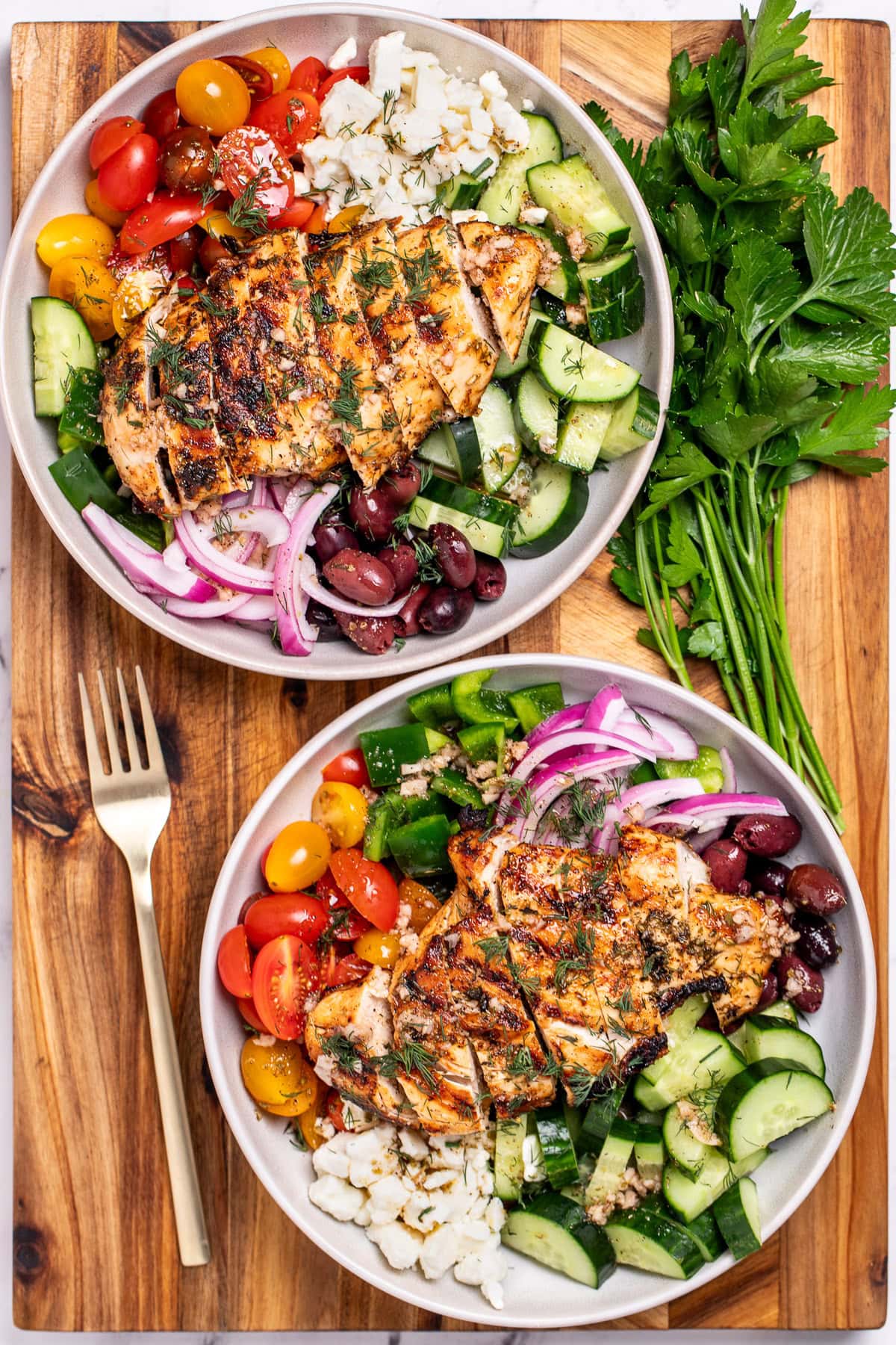 Two bowls with Greek salad and sliced grilled chicken on top, next to a fork and fresh parsley, resting on a cutting board.