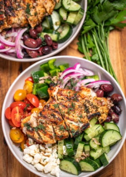 A bowl with vegetables, feta, olives, and sliced grilled chicken topped with fresh dill sitting on a cutting board, in front of another bowl and fresh parsley.