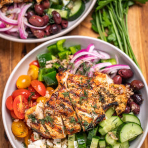A bowl with vegetables, feta, olives, and sliced grilled chicken topped with fresh dill sitting on a cutting board, in front of another bowl and fresh parsley.