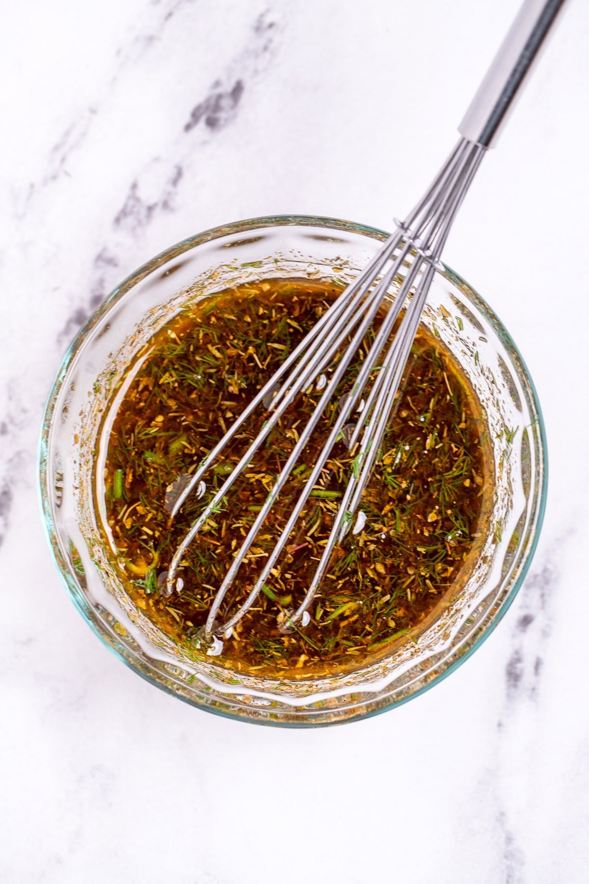 A small glass bowl on a table, full of greek marinade, with a whisk resting in it.