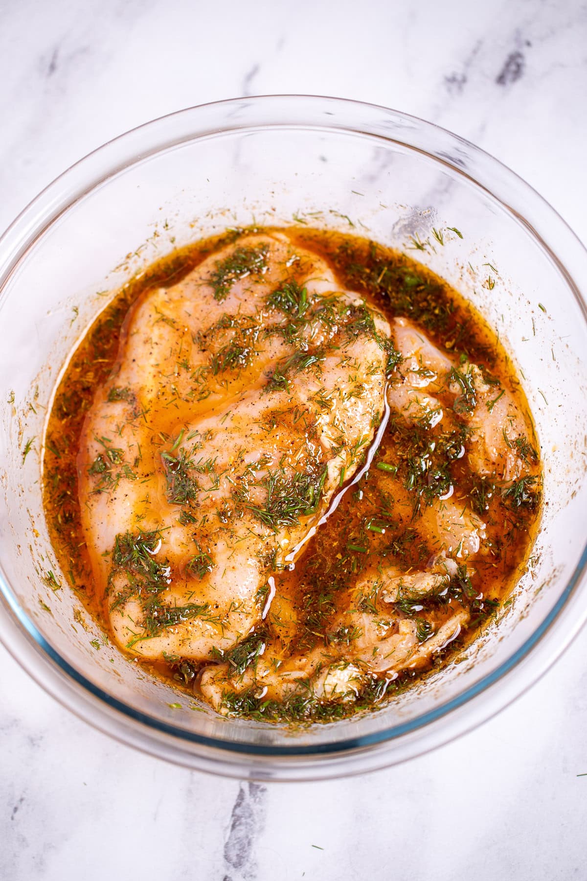 A glass bowl with chicken breast in greek marinade on a table.