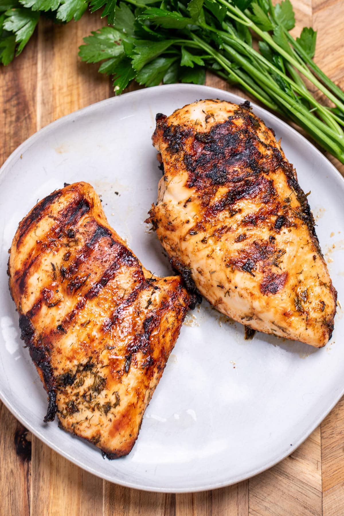 Two Greek marinated chicken breasts on a plate, resting on a cutting board next to fresh parsley.
