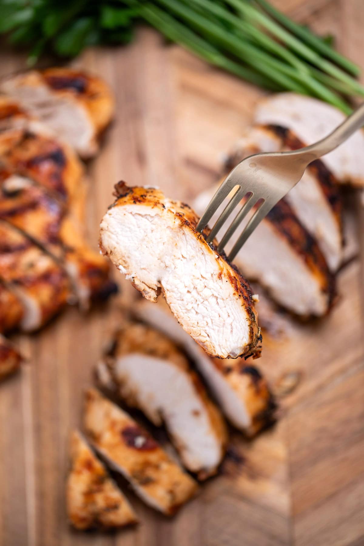 A cutting board with Greek marinated chicken sliced next to fresh parsley, and a fork lifting a piece up.