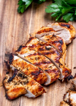A cutting board with Greek marinated chicken sliced next to fresh parsley.