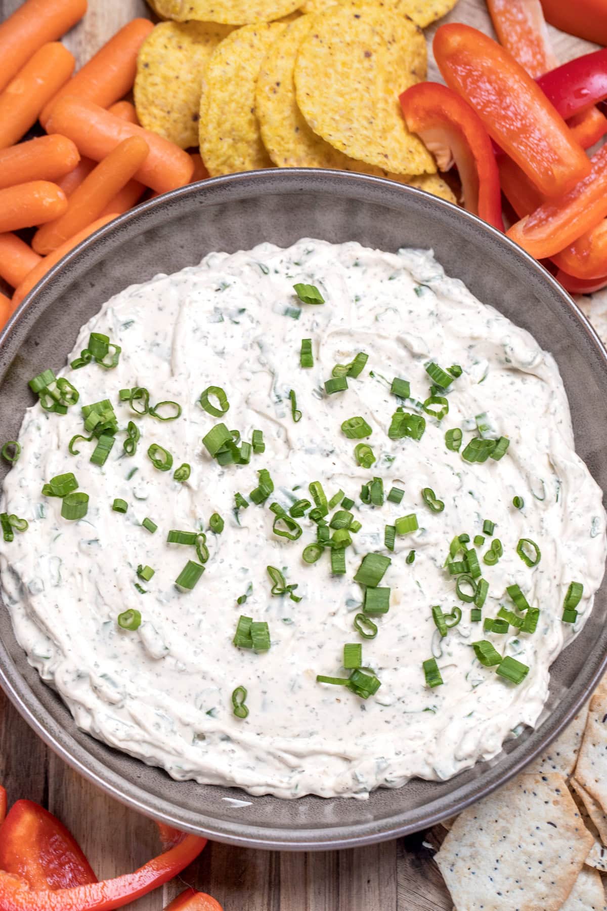 A bowl of green onion dip topped with chopped green onion, resting on a cutting board, surrounded by sliced bell pepper, crackers, baby carrots, and tortilla chips.