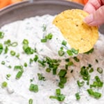 A bowl of green onion dip topped with chopped green onion, resting on a cutting board, with a hand dipping a tortilla chip into it.