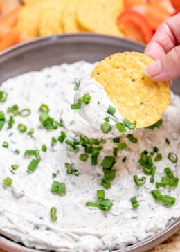 A bowl of green onion dip topped with chopped green onion, resting on a cutting board, with a hand dipping a tortilla chip into it.