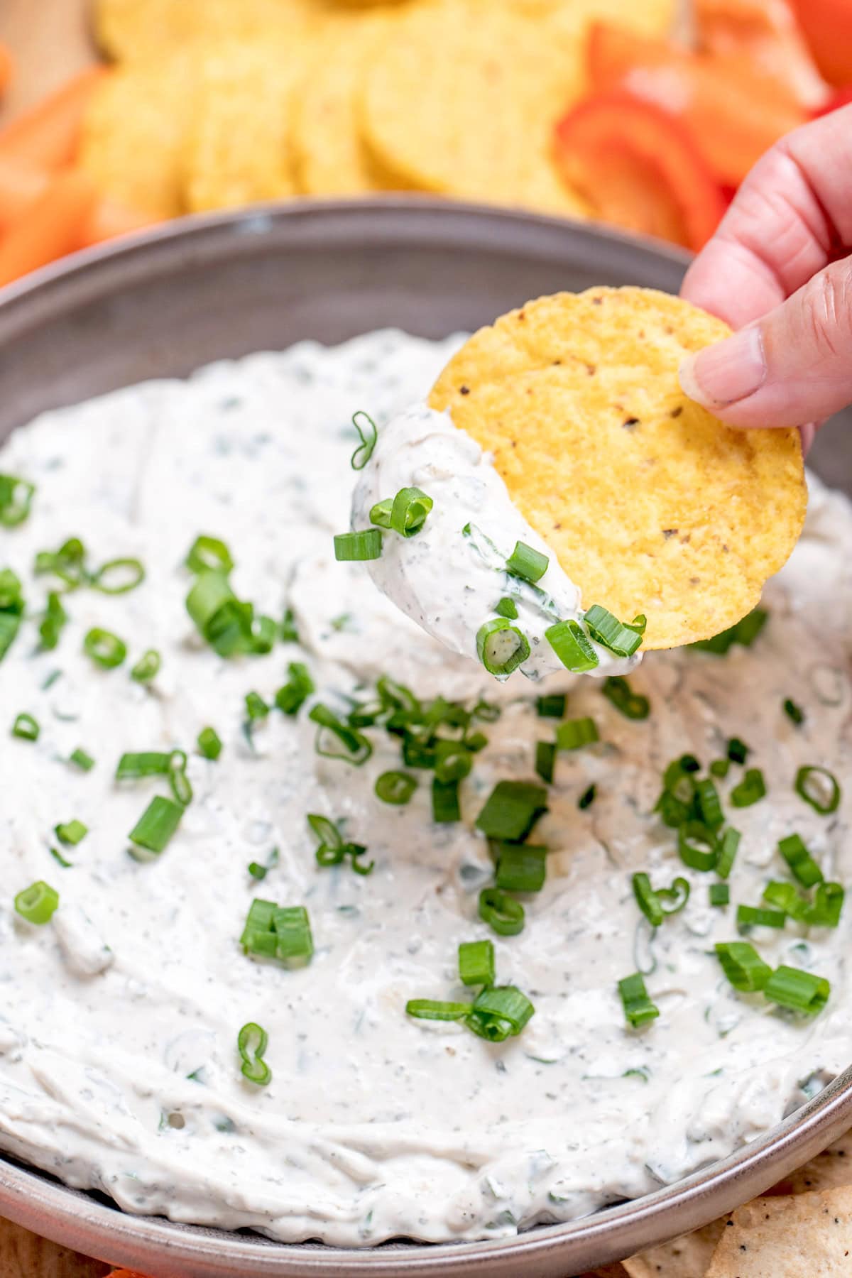 A bowl of green onion dip topped with chopped green onion, resting on a cutting board, with a hand dipping a tortilla chip into it.