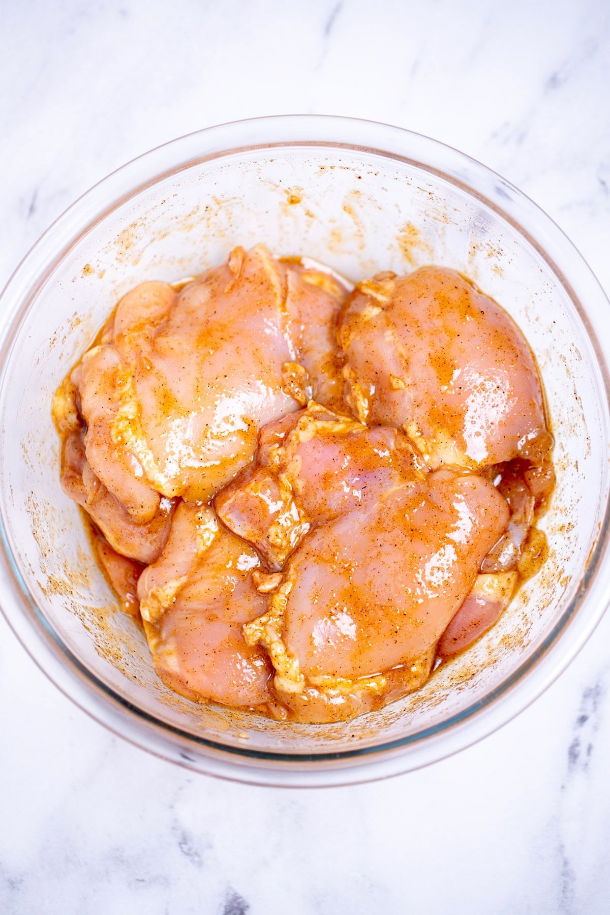 A mixing bowl with chicken thighs covered in marinade, on a table.