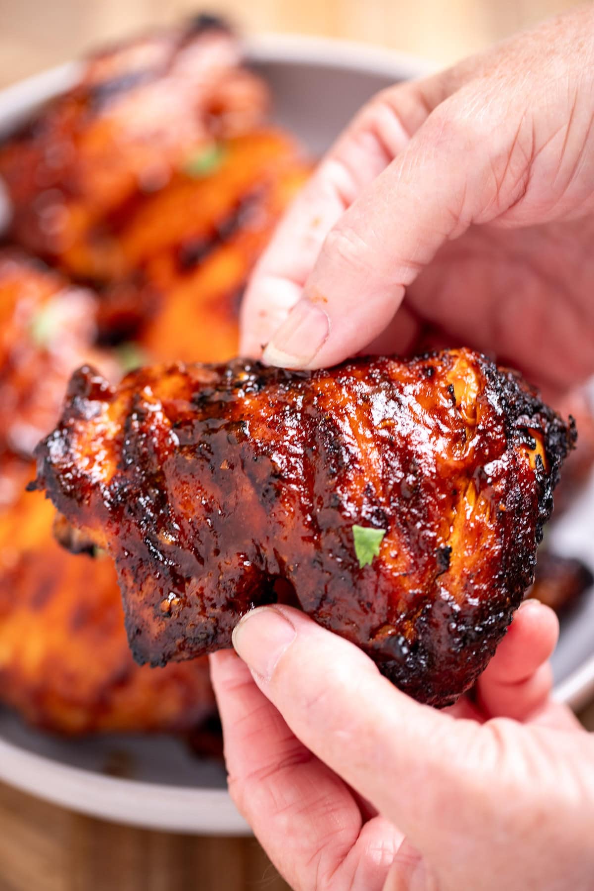 Two hands holding a chicken thigh covered in barbecue sauce, in front of a bowl of thighs.